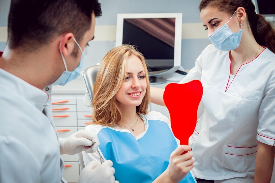 Patient smiling while holding a red heart-shaped mirror, with dental professionals in masks discussing restorative dental treatments in a modern clinic setting.