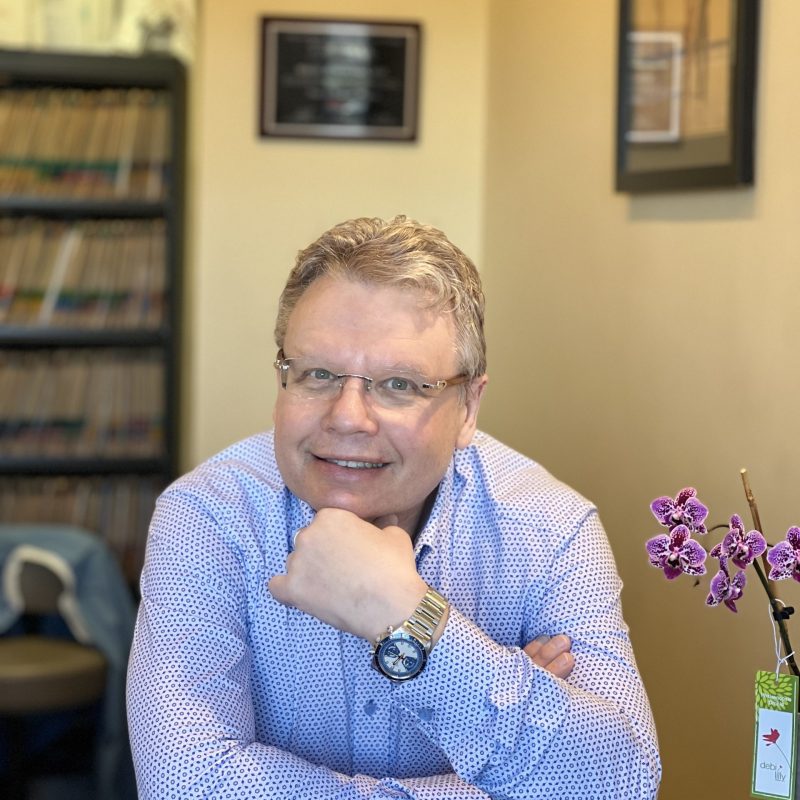 Dr. Sergey Zemlyansky at Harborside Dental, smiling in office setting with dental records in background and orchid plant.